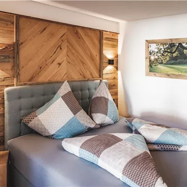 A modern bedroom with a cozy bed and colorful cushions. On the wall, there is a wooden panel and a landscape picture.