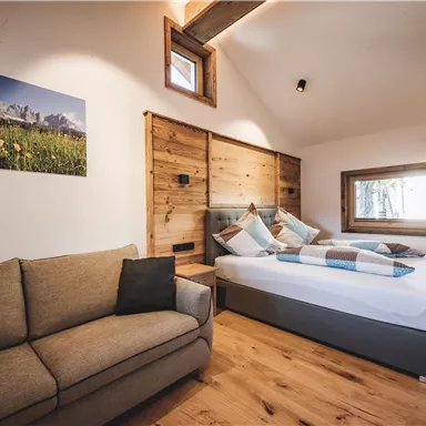 A modern bedroom with a double bed and a cozy sofa. Wooden paneling and large windows create a bright, inviting atmosphere.