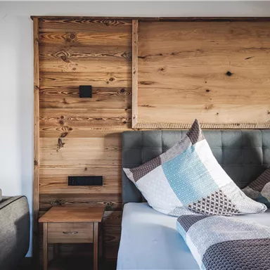 A cozy bedroom with a wooden panel headboard and decorative pillows. On the left is a gray sofa and a nightstand.