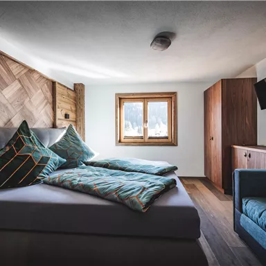 A modern bedroom with a cozy bed, colorful cushions, and a small sofa. A window with a view outside provides daylight.