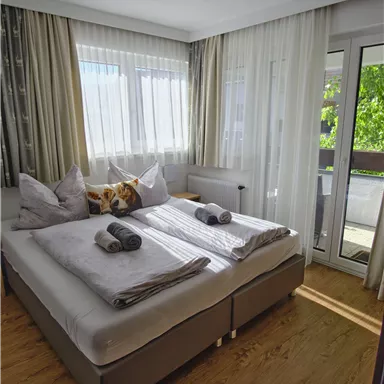 Ein helles Schlafzimmer mit einem Doppelbett und zwei Kissen. Große Fenster sorgen für viel Tageslicht und bieten Zugang zu einem Balkon.