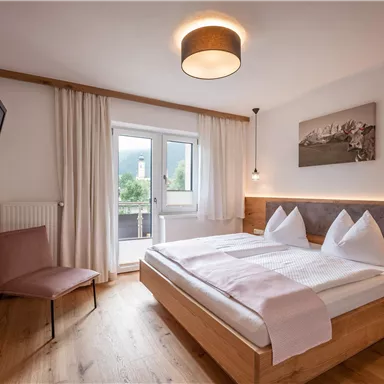 A modern bedroom with a double bed, wooden furniture, and a TV. Large windows provide plenty of daylight and a view outside.