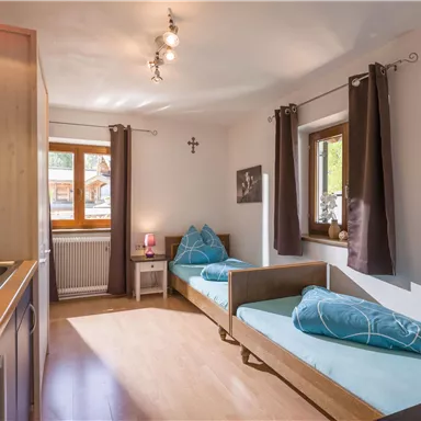 Ein gemütliches Zimmer mit zwei Einzelbetten und blauen Kissen. Die Fenster bieten Tageslicht und die Küche ist praktisch integriert.