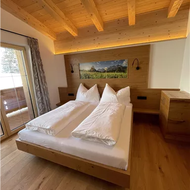 Ein gemütliches Schlafzimmer mit einem Holzbett und zwei großen Kissen. Der Raum hat große Fenster und einen warmen Holzlook.