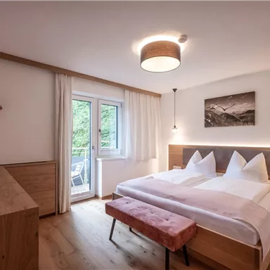 A modern bedroom with a large double bed and wooden decor. A window opens up the view outside, and a cozy seating area invites relaxation.