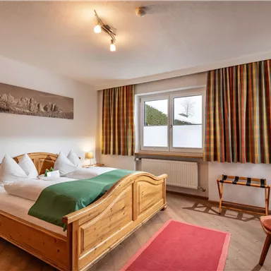 A cozy bedroom with a wooden double bed and colorful curtains. The room has a window with an outside view and a pleasant atmosphere.