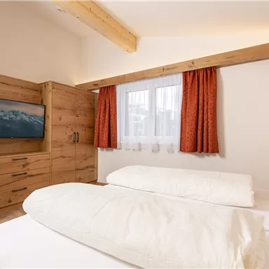 Ein gemütliches Schlafzimmer mit zwei Betten und warmen Holzmöbeln. Große Fenster und dekorative Vorhänge schaffen eine einladende Atmosphäre.