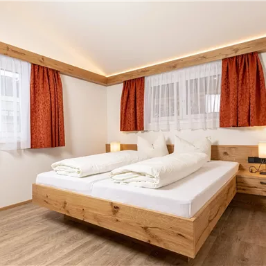 Ein helles Schlafzimmer mit einem großen Doppelbett und Holzdetails. Die Fenster sind mit weißen Vorhängen und roten Gardinen dekoriert.