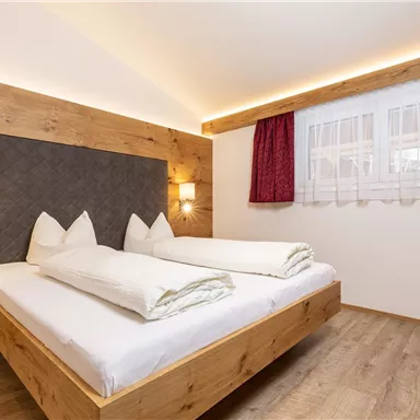 Ein gemütliches Schlafzimmer mit einem Doppelbett aus Holz. Die Wände sind hell, und das Zimmer hat große Fenster mit Vorhängen in Rot.
