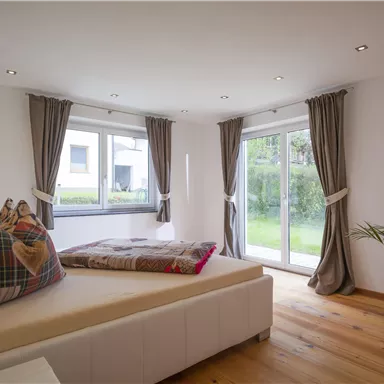 Ein modernes Schlafzimmer mit einem großen Bett und gemütlichen Kissen. Große Fenster lassen viel Licht herein und bieten einen Blick auf den Garten.