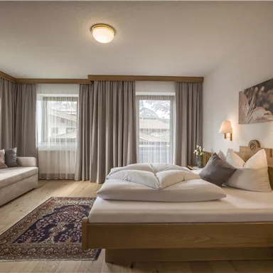 A bright bedroom with a double bed, a cozy day bed, and large windows. The walls are in neutral colors, with curtains and pleasant lighting.