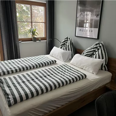 A cozy bedroom with a double bed and striped bedcovers. Large windows provide plenty of daylight and a view of the outdoors.