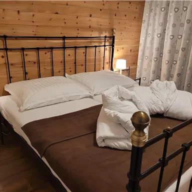 Ein gemütliches Schlafzimmer mit einem Metallbett und weißen Kissen. An den Wänden sind Holzpaneele und die Fenster sind mit Vorhängen dekoriert.