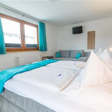 Ein gemütliches Schlafzimmer mit einem großen Bett und hellen Kissen. Die Fenster sind mit türkisen Vorhängen dekoriert und eine Ruhesitzecke ist vorhanden.