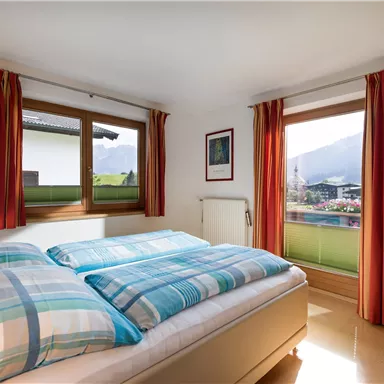 Ein helles Schlafzimmer mit einem Doppelbett und bunten Vorhängen. Durch die Fenster ist eine schöne Aussicht auf die Berge zu sehen.