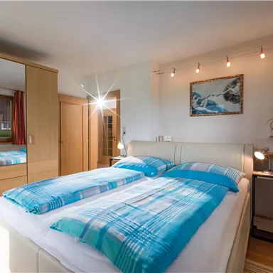 Ein gemütliches Schlafzimmer mit einem Doppelbett und blauen Bettwäsche. Es gibt einen großen Spiegel und Fenster, die viel Licht hereinlassen.