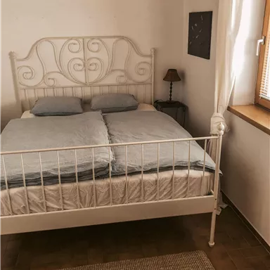 Ein gemütliches Schlafzimmer mit einem weißen Metallbett und blauen Bettdecken. Neben dem Bett steht eine kleine Lampe und ein Fenster mit Tageslicht.