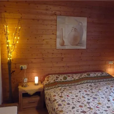 A cozy bedroom with wooden walls and a bed adorned with a colorful blanket. There are candles on the nightstands and gentle lighting from a decorative tree.