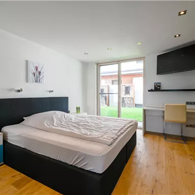 Ein modernes Schlafzimmer mit einem großen Bett und Holzboden. Große Fenster lassen viel Tageslicht herein und bieten Zugang zu einem kleinen Außenbereich.