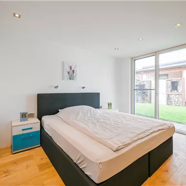 Ein modernes Schlafzimmer mit einem großen Bett und hellen Wänden. Große Fenster lassen viel Licht herein und bieten Blick auf einen Garten.