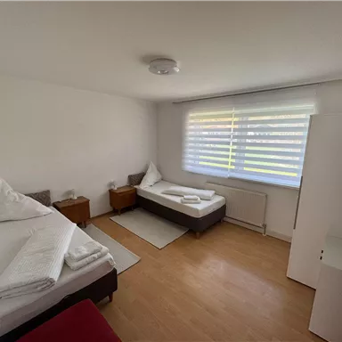Ein einfaches Zimmer mit zwei Einzelbetten und minimalistischer Einrichtung. Es gibt einen hellen Holzboden und viel natürliches Licht durch das Fenster.