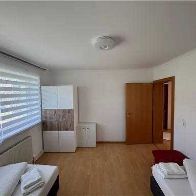 Ein einfaches und helles Schlafzimmer mit zwei Betten und Holzfußboden. Es gibt einen Schrank und einen weiteren Raum im Hintergrund.