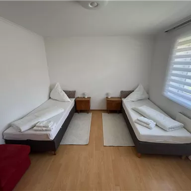 Ein helles Zimmer mit zwei Einzelbetten und Nachttischen. Die Wände sind weiß und es gibt große Fenster mit Jalousien.