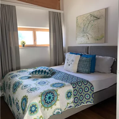 A cozy bedroom with a double bed and colorful cushions. The walls are light, and a window provides ample daylight.
