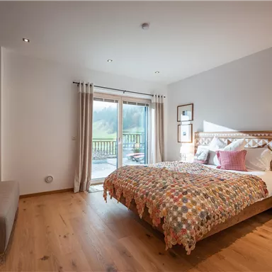 Ein gemütliches Schlafzimmer mit einem großen Doppelbett und einer bunten Decke. Große Fenster ermöglichen einen Blick auf die Natur draußen.