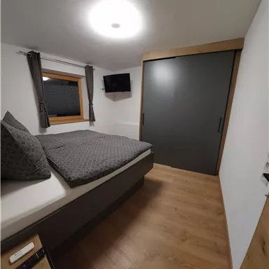 A modern bedroom with a large bed and gray bedding. The room features a television and a built-in wardrobe.