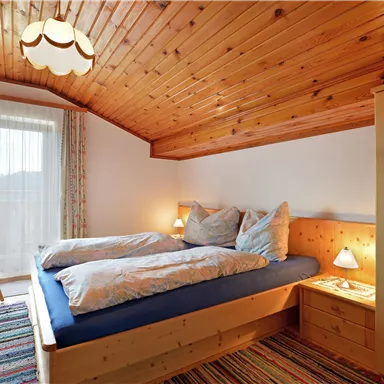 Ein gemütliches Schlafzimmer mit Holzdecke und zwei Betten. Neben dem Bett befinden sich eine Nachtlampe und ein Stuhl, sowie ein Fenster mit Blick nach draußen.