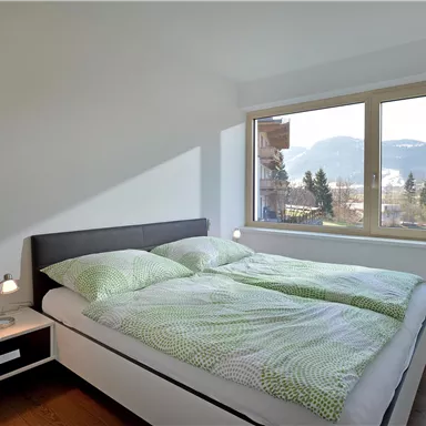 A modern bedroom with a double bed and green bedding. Large windows offer a view of the mountains and plenty of natural light.