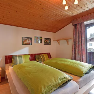A cozy bedroom with two beds and green bedcovers. There are large windows that let in plenty of light, and wooden details in the ceiling.