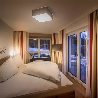 A cozy bedroom with a large bed and bright curtains. Large windows offer a view outside.