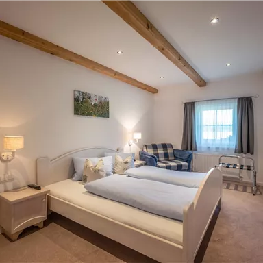 Ein gemütliches Schlafzimmer mit einem Doppelbett und einem zusätzlichen Bett. Der Raum hat Holzdeckenbalken, eine gemütliche Sitzgelegenheit und ein Fenster mit Vorhängen.