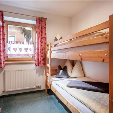 A small room with colorful curtains and a wooden bunk bed. It has a window with a view and a wardrobe in the background.