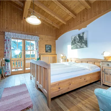 Ein gemütliches Zimmer mit Holzdecken und einem großen Betten. Es gibt einen Tisch und Stühle sowie einen kleinen Balkon mit Blick nach draußen.