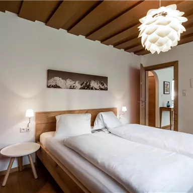 A modern bedroom with a double bed and wood decor. A stylish lamp hangs from the ceiling, and there is a door leading to another room.