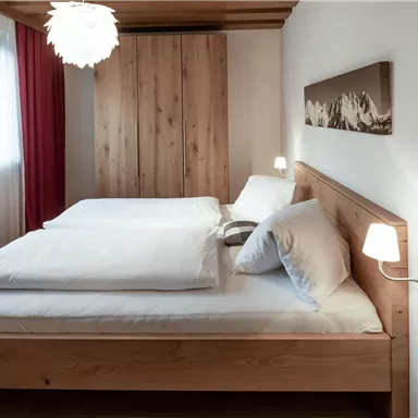 A cozy bedroom with a large wooden bed and two white comforters. The room is bright and modernly furnished, with a window that lets in daylight.