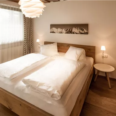 A cozy bedroom with a large double bed and wooden details. The room has windows with curtains and a modern lamp.