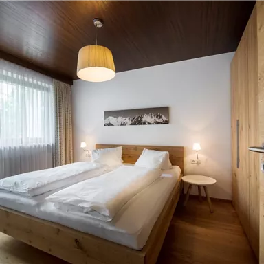 A cozy bedroom with a double bed and wooden-colored furniture. Large windows let in plenty of daylight.