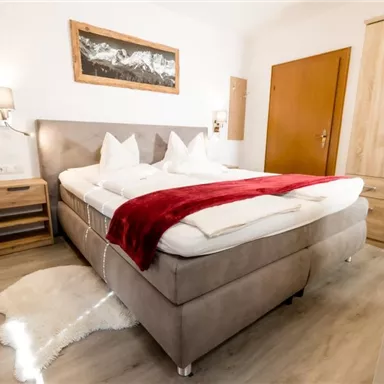 Ein modernes Schlafzimmer mit einem gemütlichen Doppelbett und roten Akzenten. Der Raum verfügt über Holz- und Textilmöbel sowie ein helles, freundliches Ambiente.