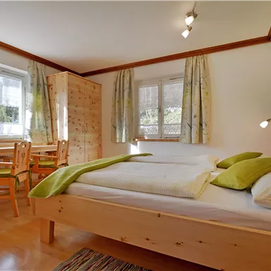 A bright bedroom with a large bed and green cushions. There is a desk and wooden shelves that create a cozy atmosphere.
