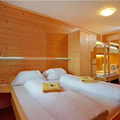 Ein gemütliches Zimmer mit Holzdekor und zwei Einzelbetten. Im Hintergrund befindet sich ein Etagenbett sowie eine warme Beleuchtung.