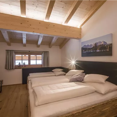 A bright and modern bedroom with a wooden beam ceiling. There are three beds, windows with curtains, and a beautiful landscape picture on the wall.