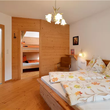 Ein gemütliches Schlafzimmer mit Holzverkleidung und einem großen Bett. Im Hintergrund befindet sich ein offenes Zimmer mit Etagenbetten.