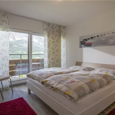 A cozy bedroom with two beds and a large window.
There are light walls, a chair, and a red carpet.