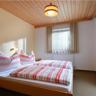 Ein gemütliches Schlafzimmer mit Holzdecke und großen Fenstern. Es gibt ein Doppelbett mit gestreiften Bettwäsche und einem Nachttisch.