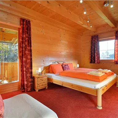 Ein gemütliches Schlafzimmer mit Holzvertäfelung und rotem Teppich. Das Bett hat orangefarbene Bettwäsche und große Fenster sorgen für viel Licht.