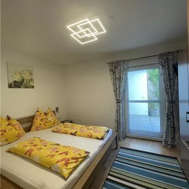 Ein helles Schlafzimmer mit einem Doppelbett und bunten Kissen. Es gibt einen Zugang zu einem Balkon und einen modernen Lichtschirm.